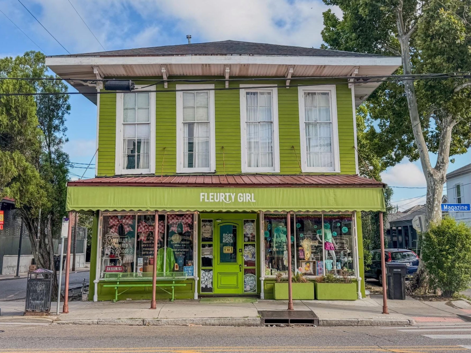A bright lime green two-story storefront building with large windows and a sign reading 'FLEURTY GIRL' above the entrance, set on a street with trees, utility poles, and a blue sky with clouds on Magazine Street in New Orleans.